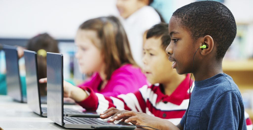 A multi-ethnic group of elementary age children are in the computer lab using laptops. A little boy is watching a video and is listening to music.